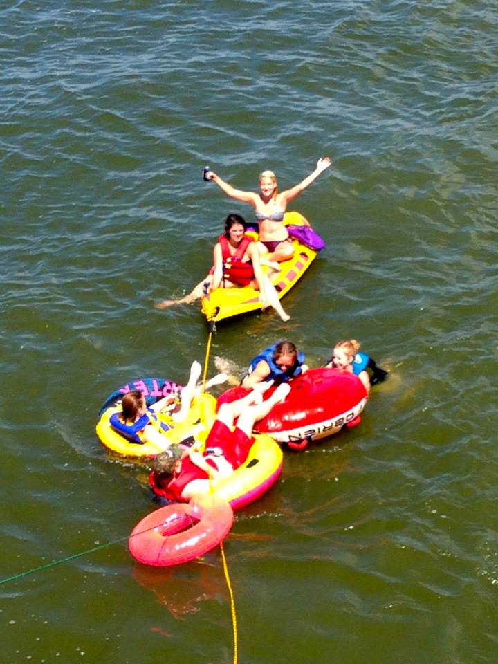 Group of kids and a woman on water floats and a yellow paddleboard with purple and yellow colors, enjoying a day on the water near the Chesapeake Bay during sunny weather.