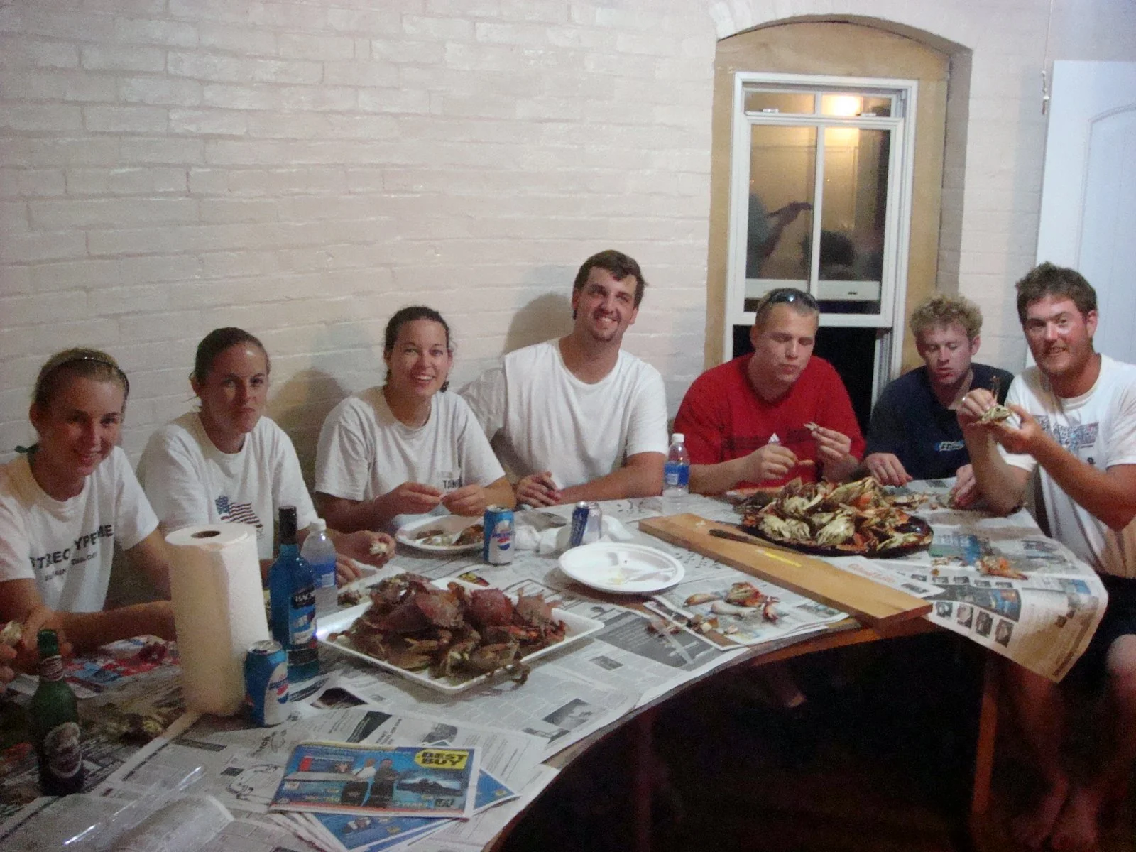 A group of seven young adults sitting around a table with seafood, drinks, and paper towels, smiling and enjoying a meal together  in the kitchen of Middle Ground Lighthouse. 