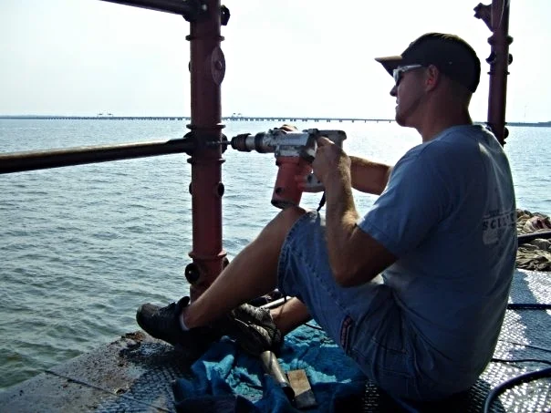 A man working on the main deck railings of Middle Ground Lighthouse, holding a power drill that is attached to a pipe.