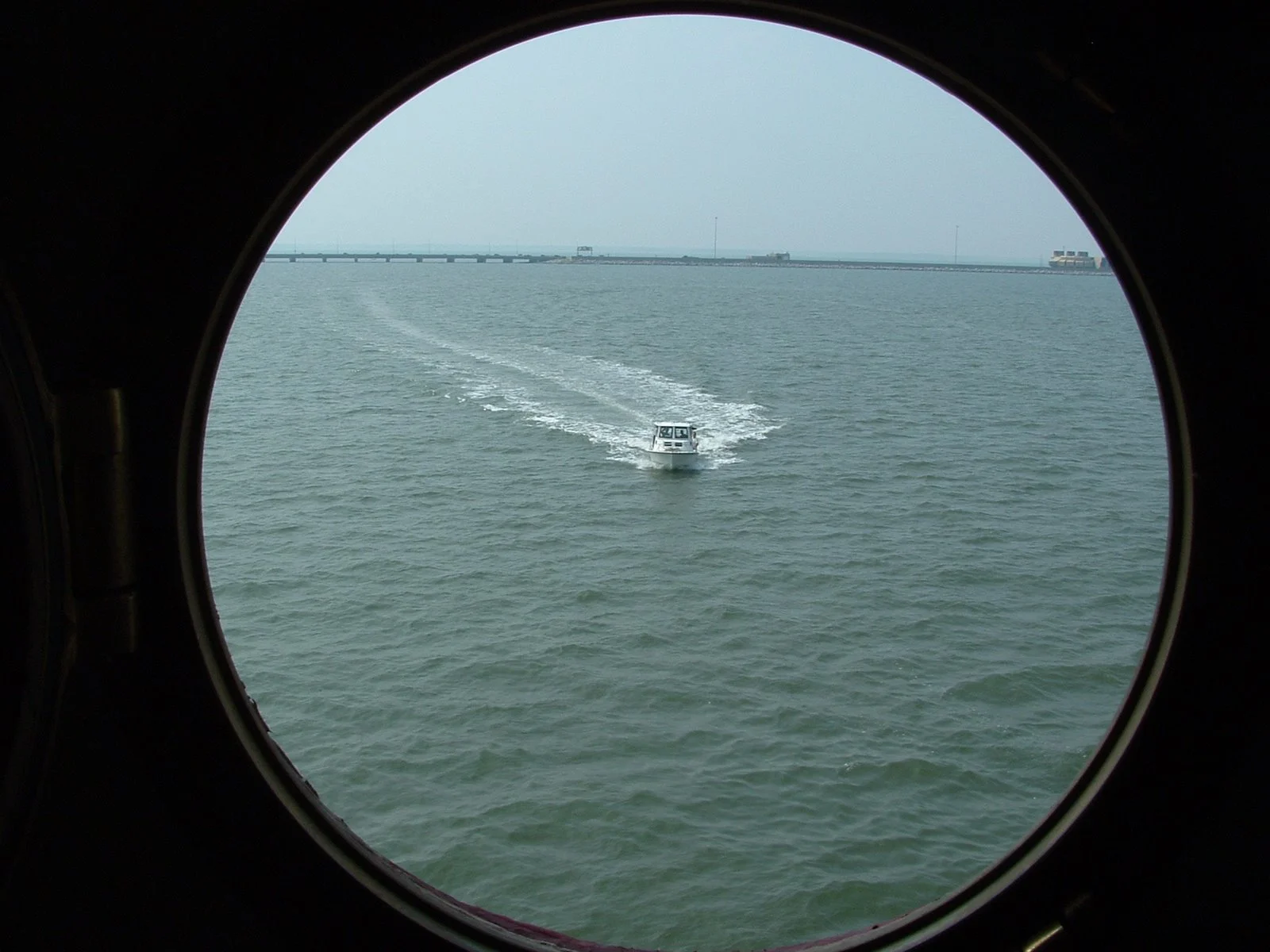 A view of a small boat sailing on the water seen through a circular porthole window on Middle Ground Lighthouse, with a bridge and distant buildings in the background.