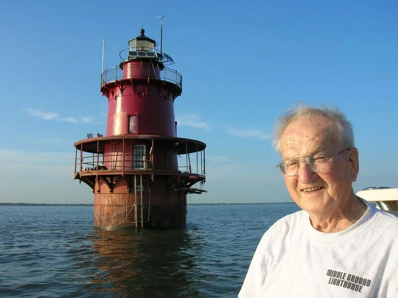 An elderly man with glasses smiling in front of Middle Ground Lighthouse in the water, under a clear blue sky.