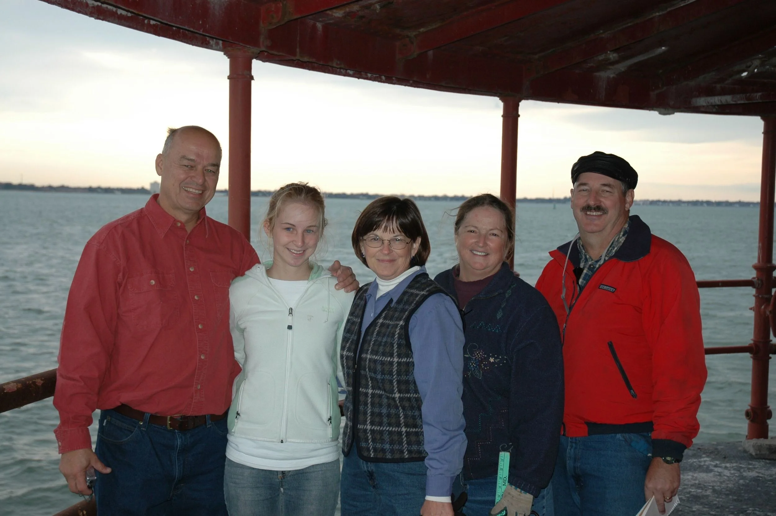 Six people standing together the main deck of Middle Ground Lighthouse near water with Hampton Roads in the background during sunset or dusk.