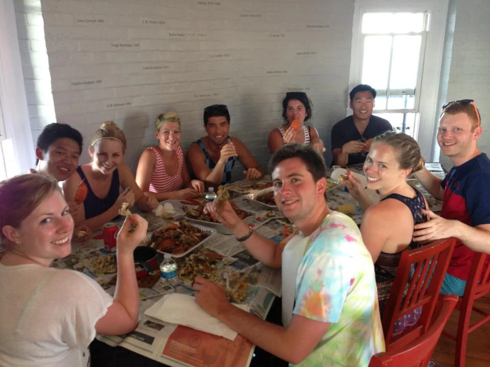 Group of eleven young adults having a meal at a table in the kitchen of Middle Ground Lighthouse, smiling and enjoying food.