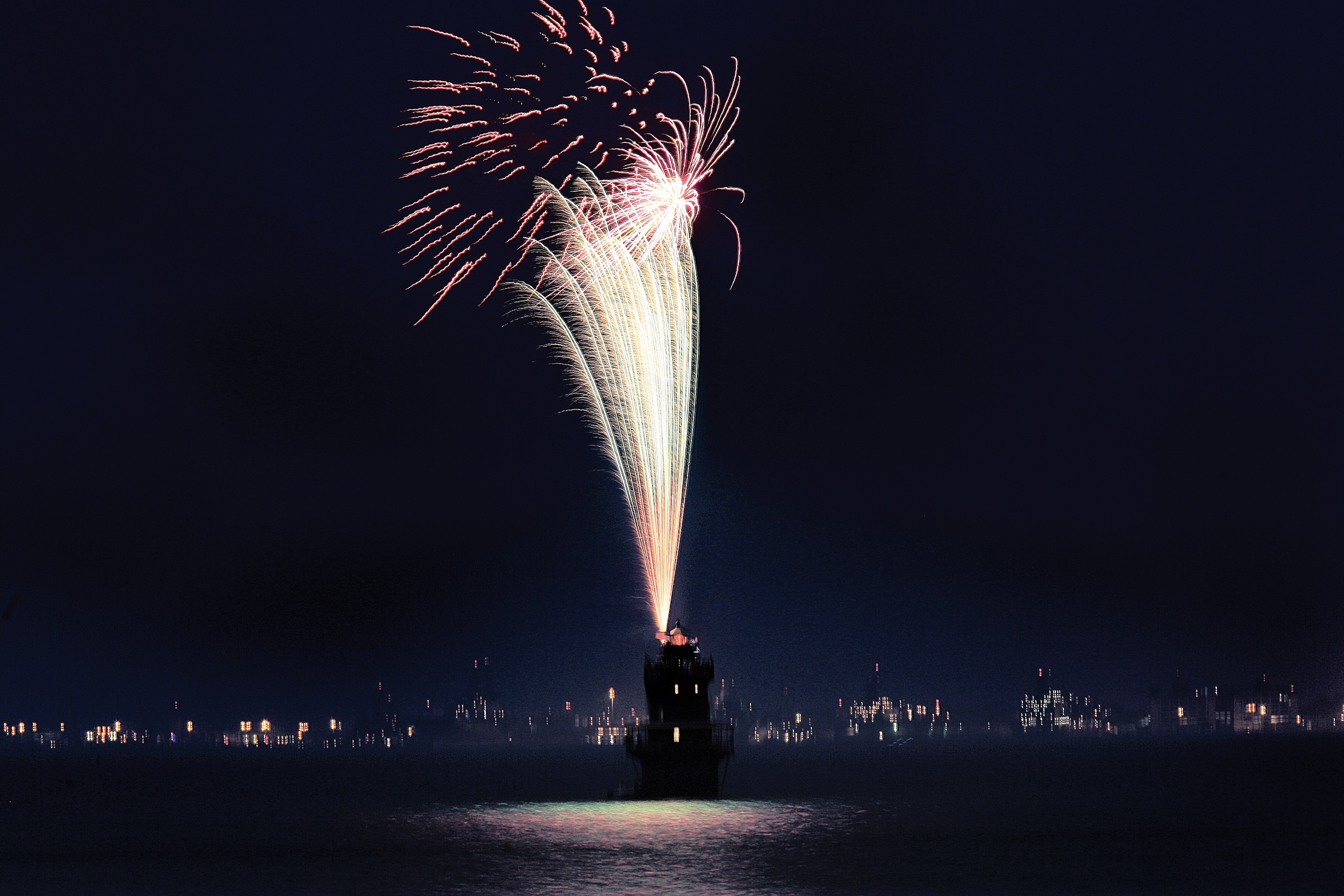 Fireworks exploding above Middle Ground Lighthouse at night, with reflections on the water, taken from the Monitor–Merrimac Memorial Bridge–Tunnel.