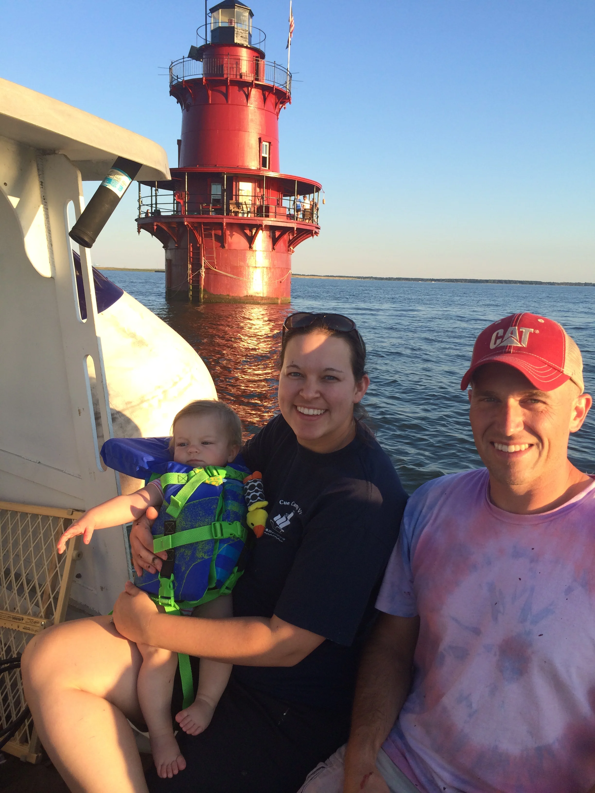 A group of people, including a woman, a man, and a baby, are on a boat with Middle Ground Lighthouse in the background on the water during sunset or late afternoon.