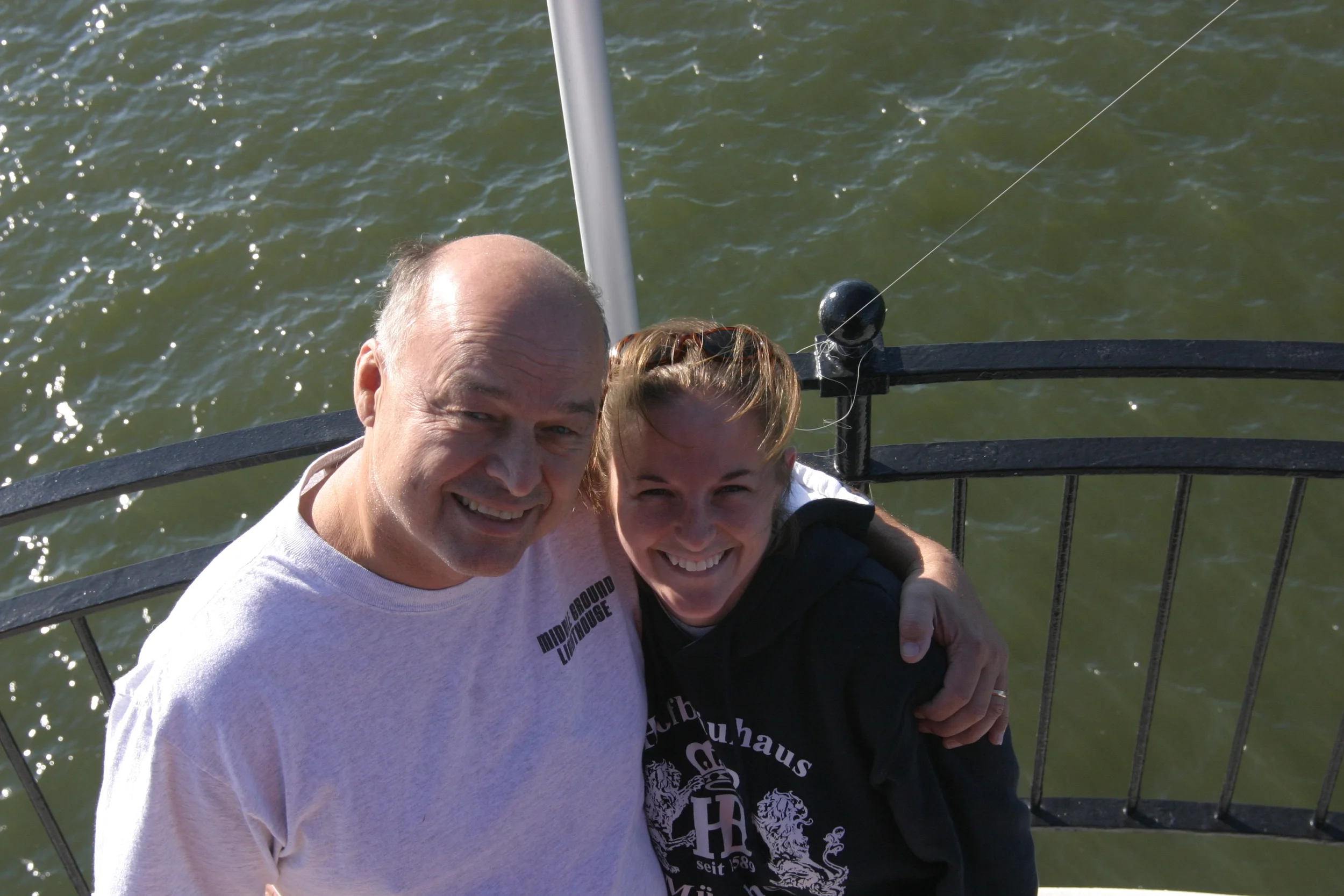 Two smiling people, an older man and a younger person, taking a selfie on Middle Ground Lighthouse with green water in the background.