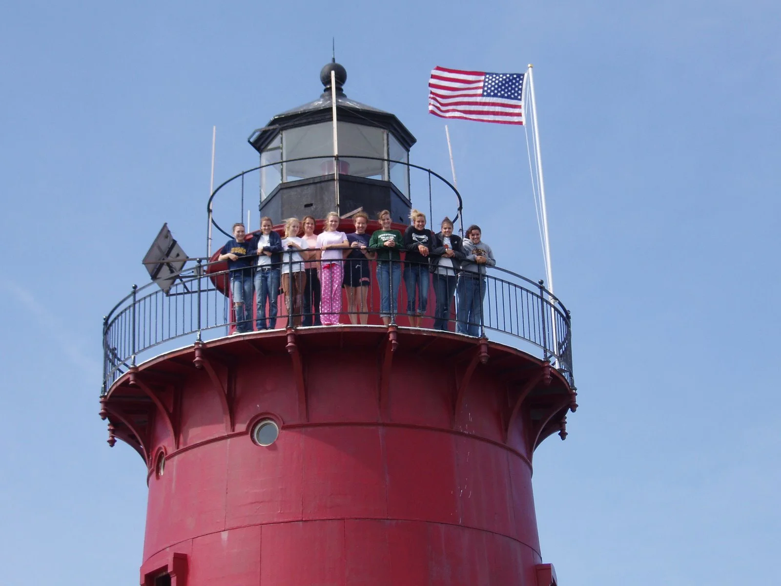 Group of nine people standing on top of Middle Ground Lighthouse with a blue sky background. An American flag is flying next to the lighthouse.