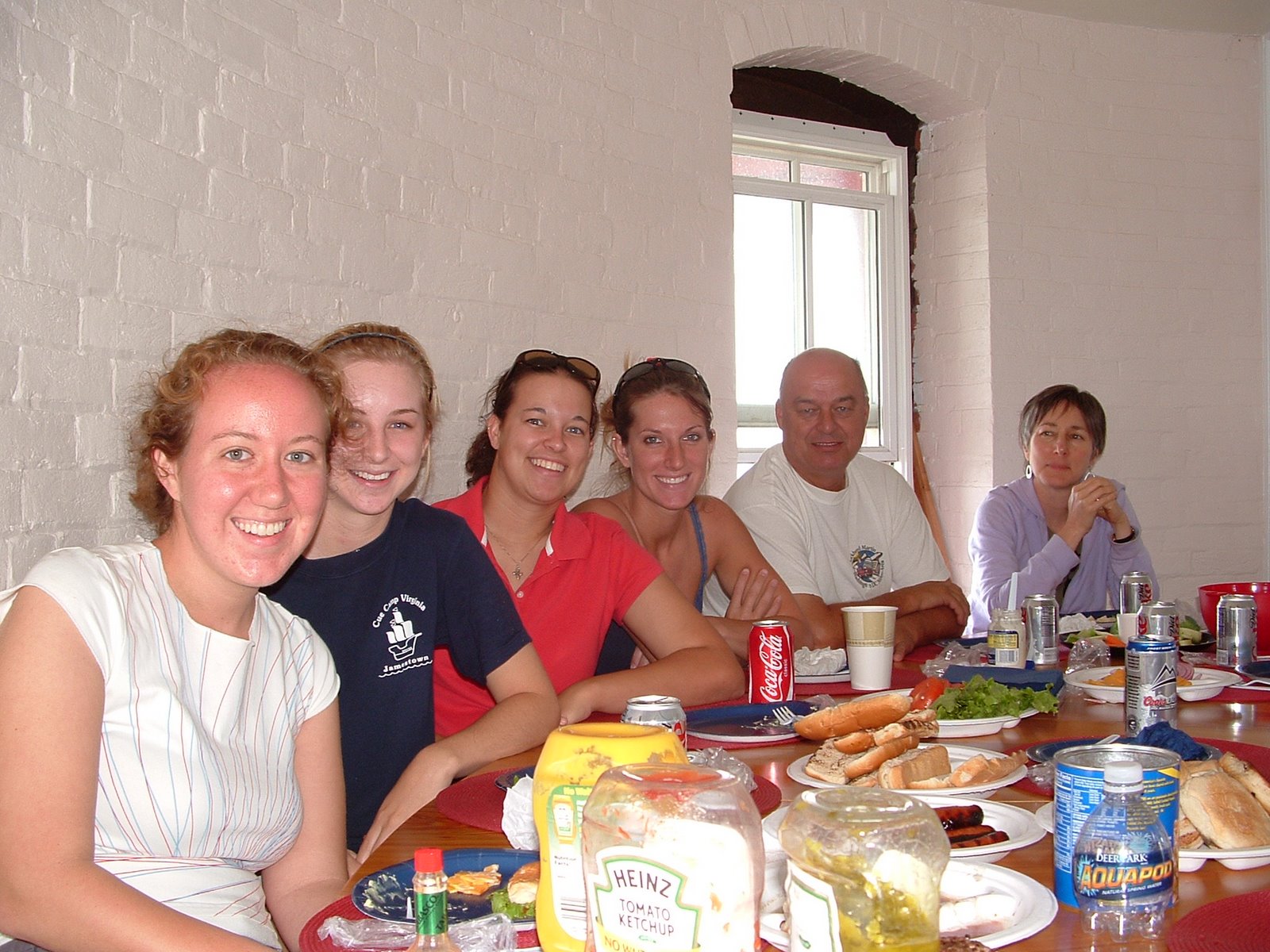 A group of six people sitting at a table, smiling, with food and drinks on the table, in the kitchen of Middle Ground Lighthouse.