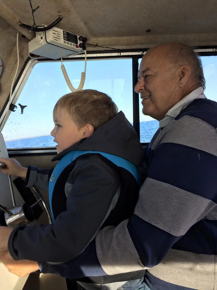 An elderly man and a young boy sitting in the cockpit of a boat, with the boy steering and the man smiling, both looking outward at the water through the boat's window.