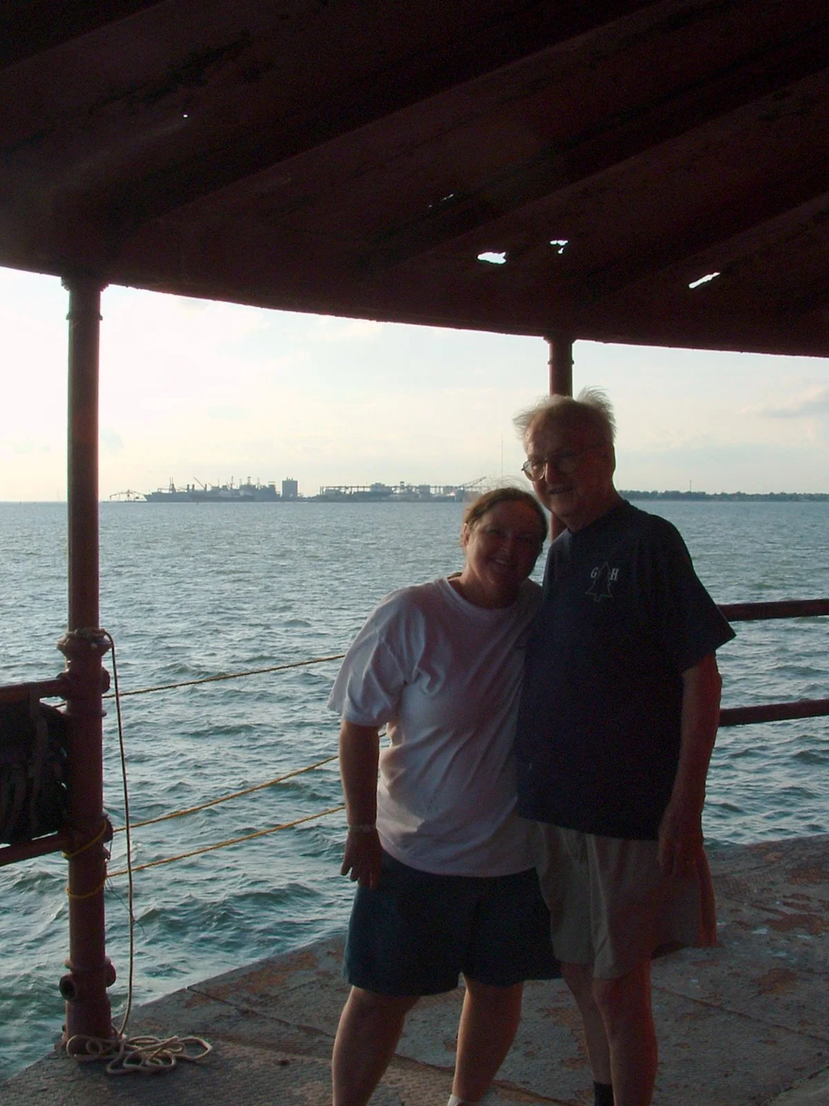 A woman and man standing on the main deck of Middle Ground Lighthouse, Hampton Roads in the background, enjoying a sunset or late afternoon by the water.