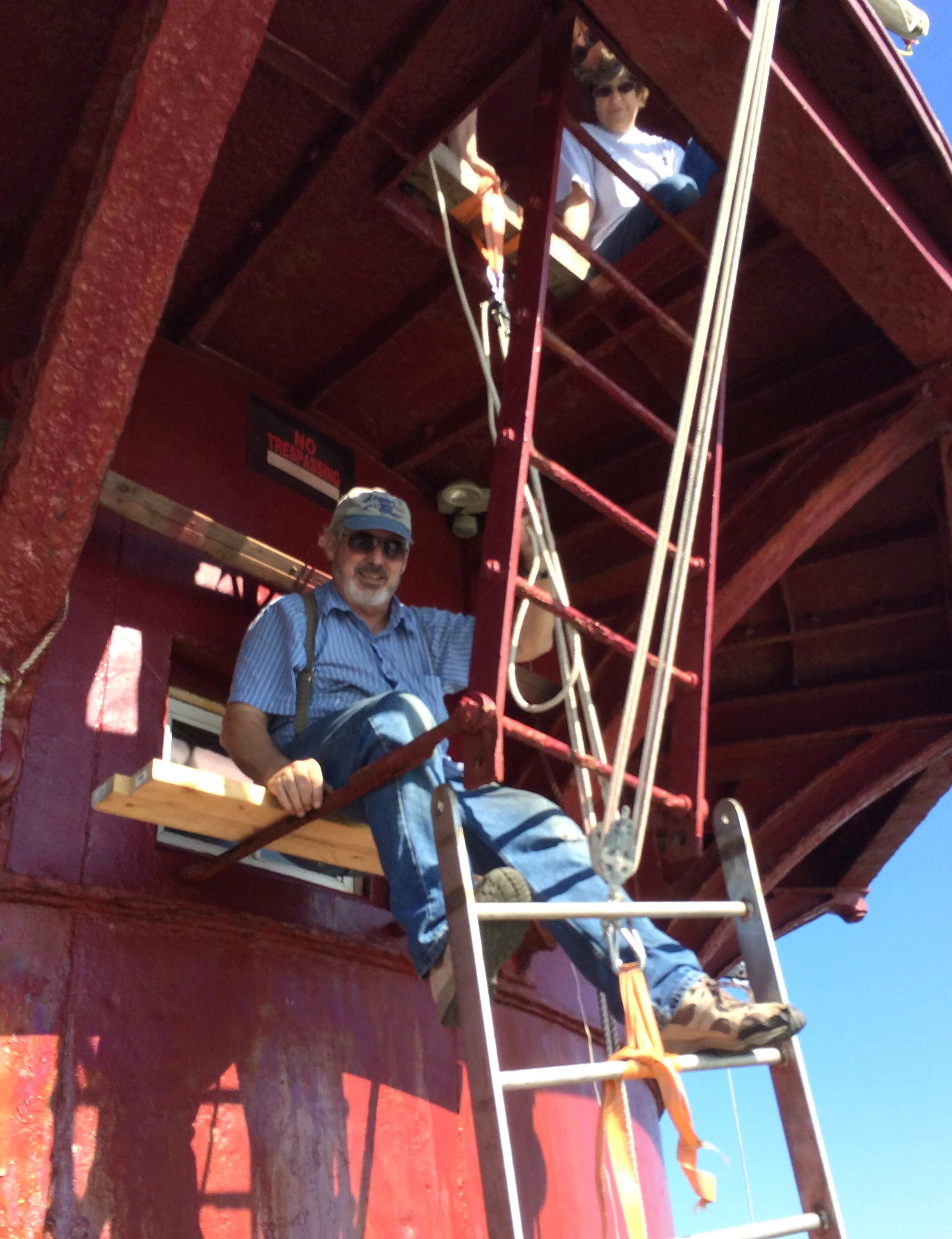 A man sitting on a red ladder platform of Middle Ground Lighthouse. The platform has a railing and is attached to a red painted structure, with a sign that says 'NO TRESPASSING' in the background. The sky is clear and blue.