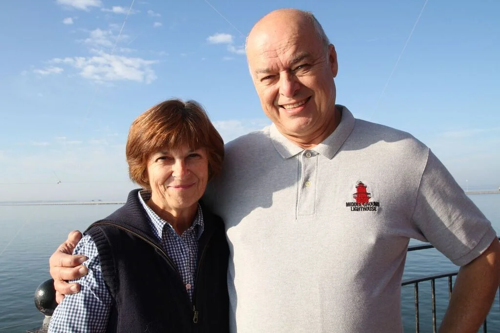A smiling older man and woman standing close together outdoors by a body of water under a clear blue sky at the top of Middle Ground Lighthouse. 