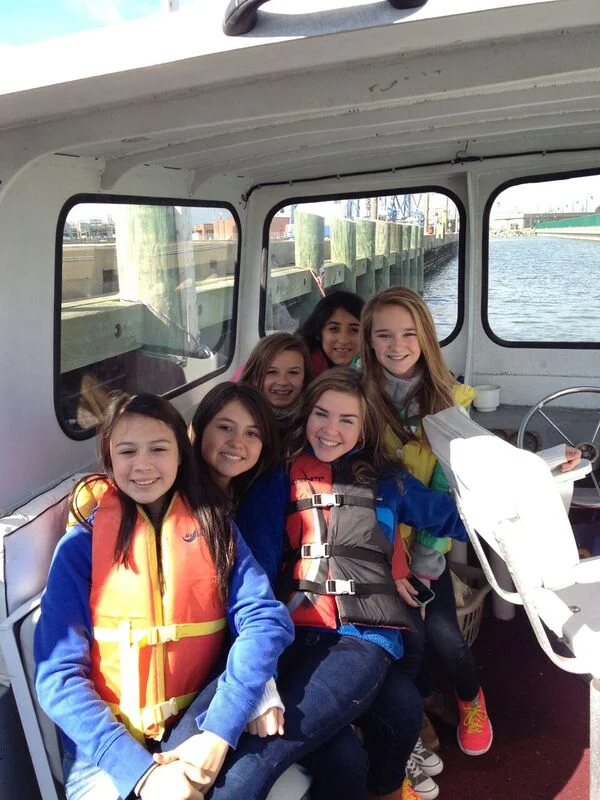 Six young girls smiling inside a boat with life jackets on, near water, possibly at a dock or marina.