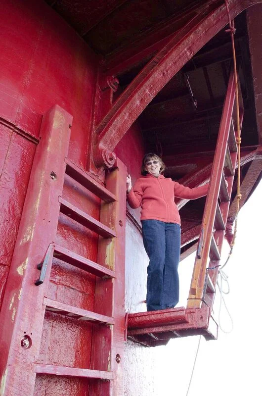 Woman standing on a small red platform at the top of Middle Ground Lighthouse, wearing a red hoodie and blue jeans, waving and smiling.