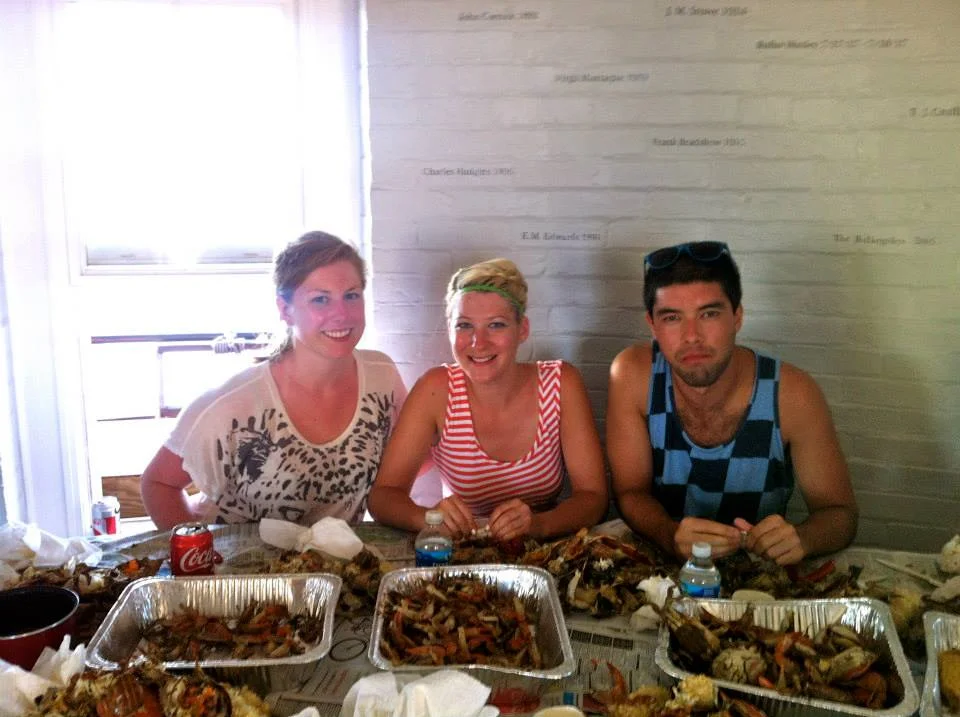 Three people sitting at a table with trays of cooked crab, bottles of water, and a can of Coca-Cola in the bright kitchen of Middle Ground Lighthouse. 