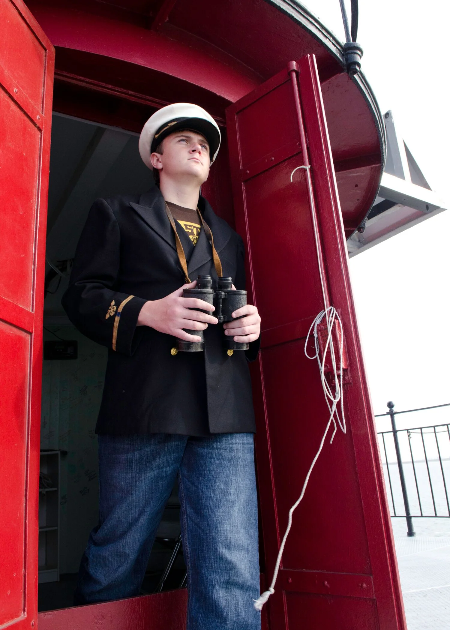 A young man dressed as a sailor looking to the distance while standing on Middle Ground Lighthouse, holding binoculars, with a white sailor hat, black blazer, and jeans.