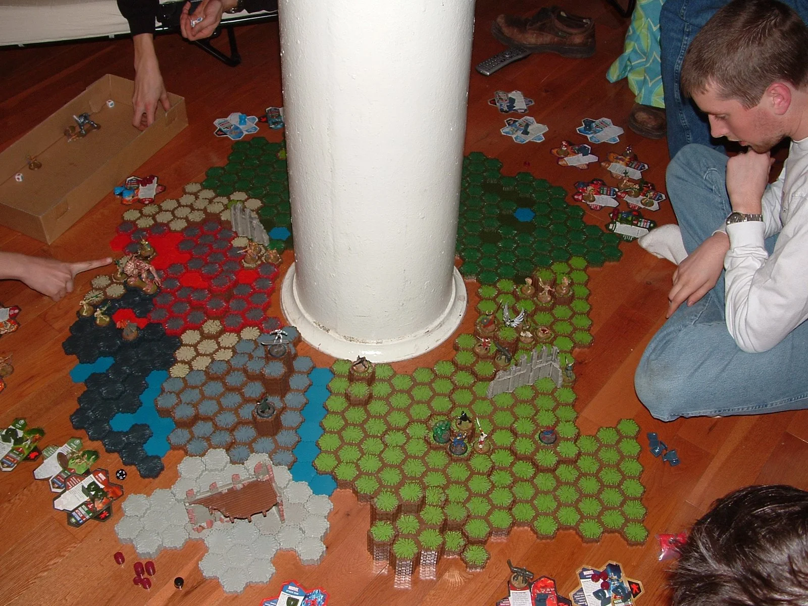 A group of people playing a tabletop strategy game on the floor, with hexagonal tiles and miniature figures arranged around a white pillar on Middle Ground Lighthouse. 