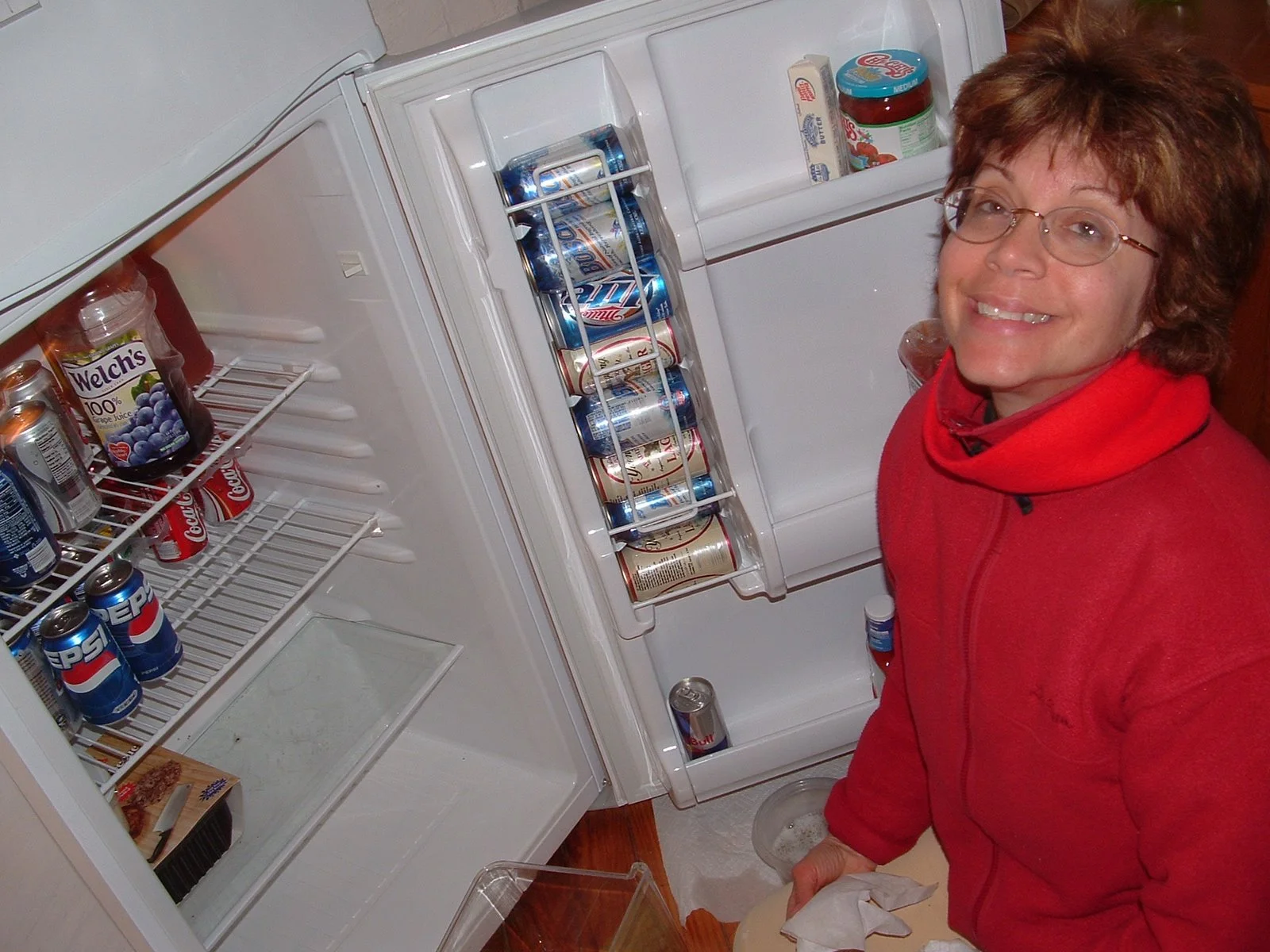 A woman in a red hoodie smiling while sitting next to an open refrigerator filled with cans of soda, yogurt, and other food items in the kitchen of Middle Ground Lighthouse. 