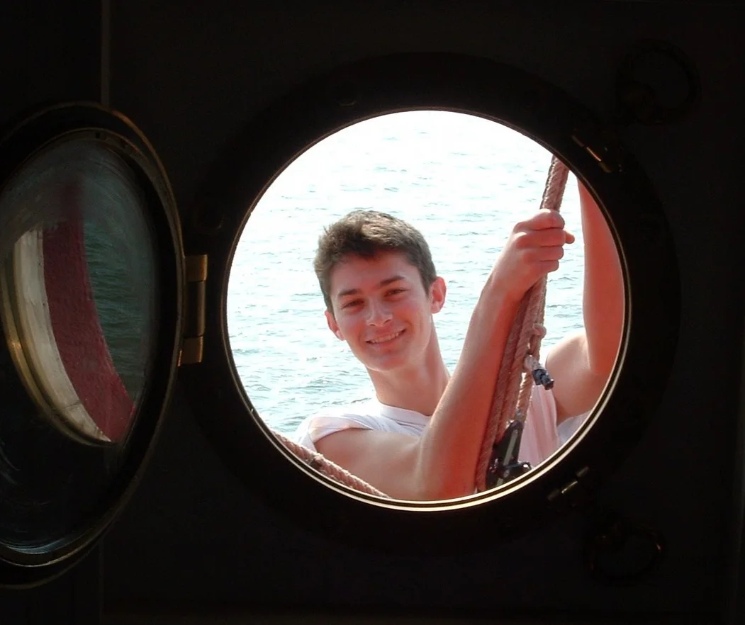 A young man smiling while working outside of an open porthole on Middle Ground Lighthouse. 
