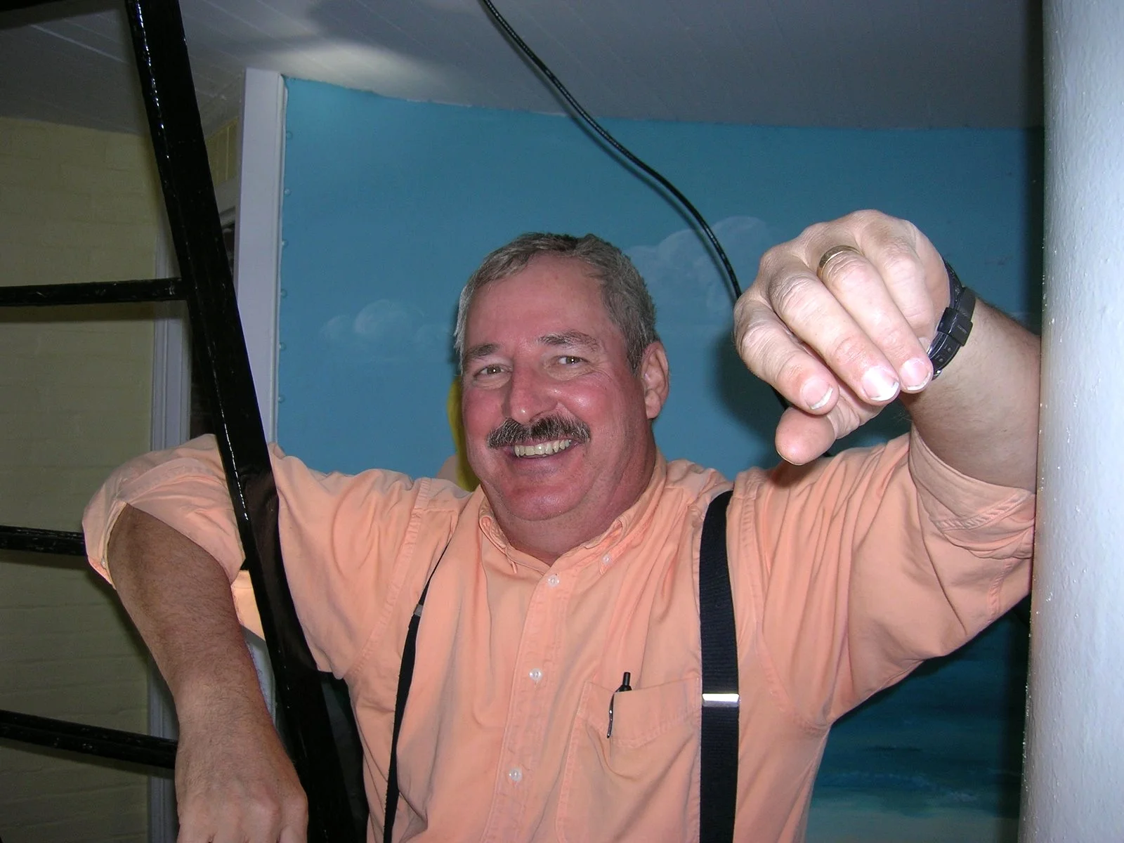 A man with gray hair, a mustache, and a big smile, leans against the ladder in the porthole room of Middle Ground Lighthouse. 