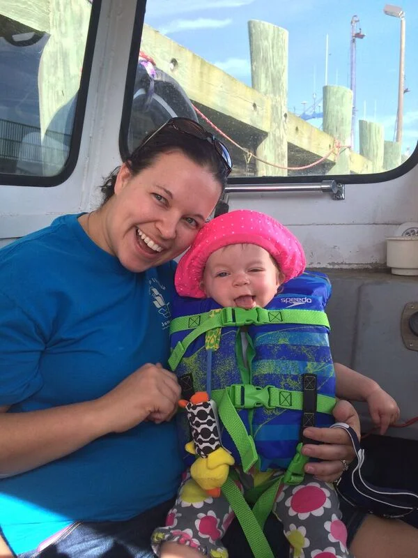 A woman and a young girl sitting inside a boat, smiling at the camera. The girl is wearing a bright pink wide-brim hat and a blue life jacket, with colorful floral pants.