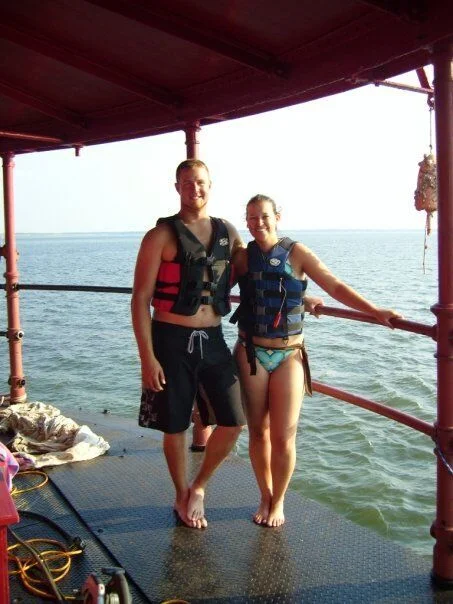 A young man and woman in life jackets standing on the main deck of Middle Ground Lighthouse by the water, smiling.