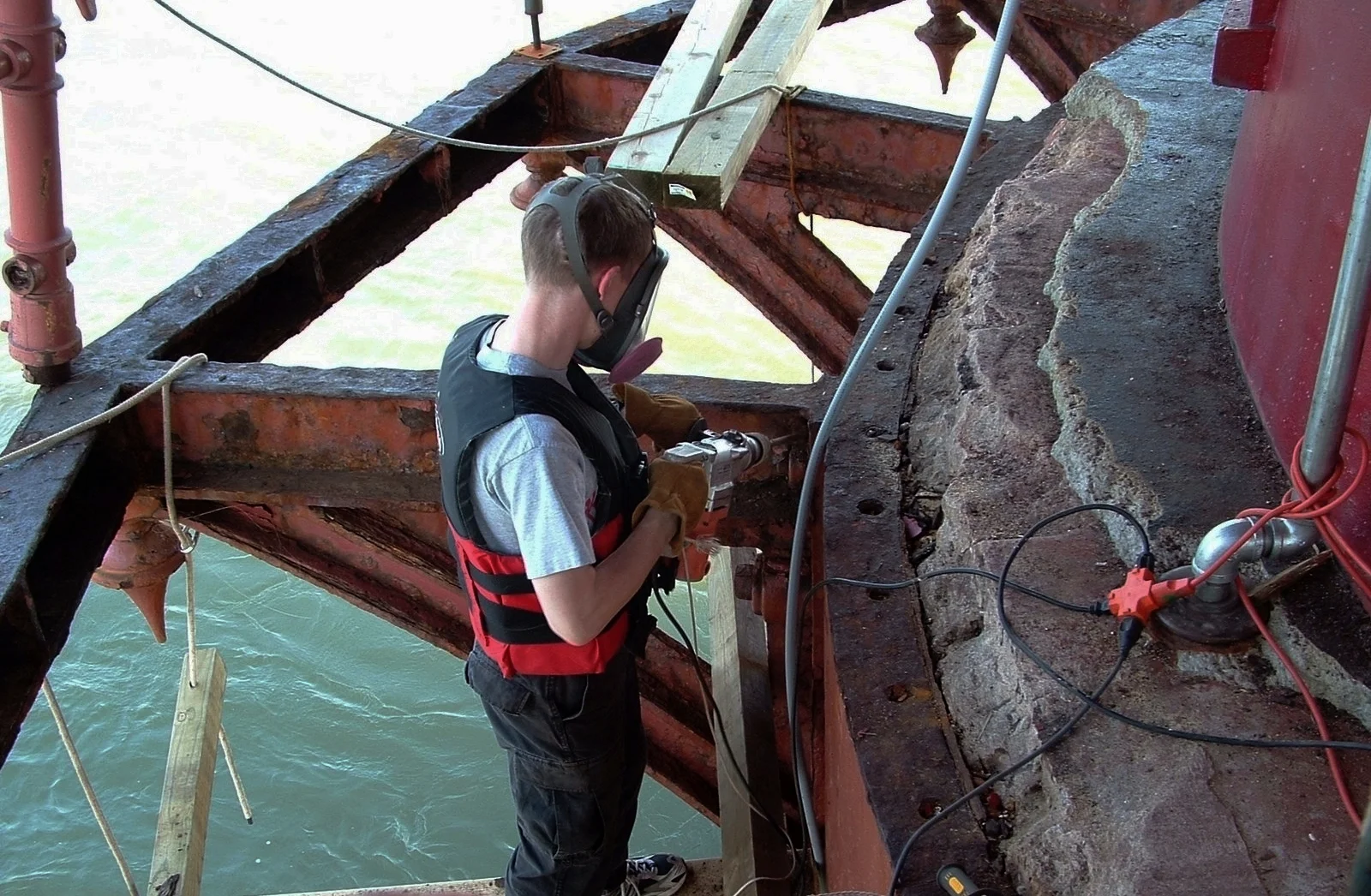 A person wearing safety gear, including a protective mask, working on a large metal and stone structure on a dock, with water in the background.