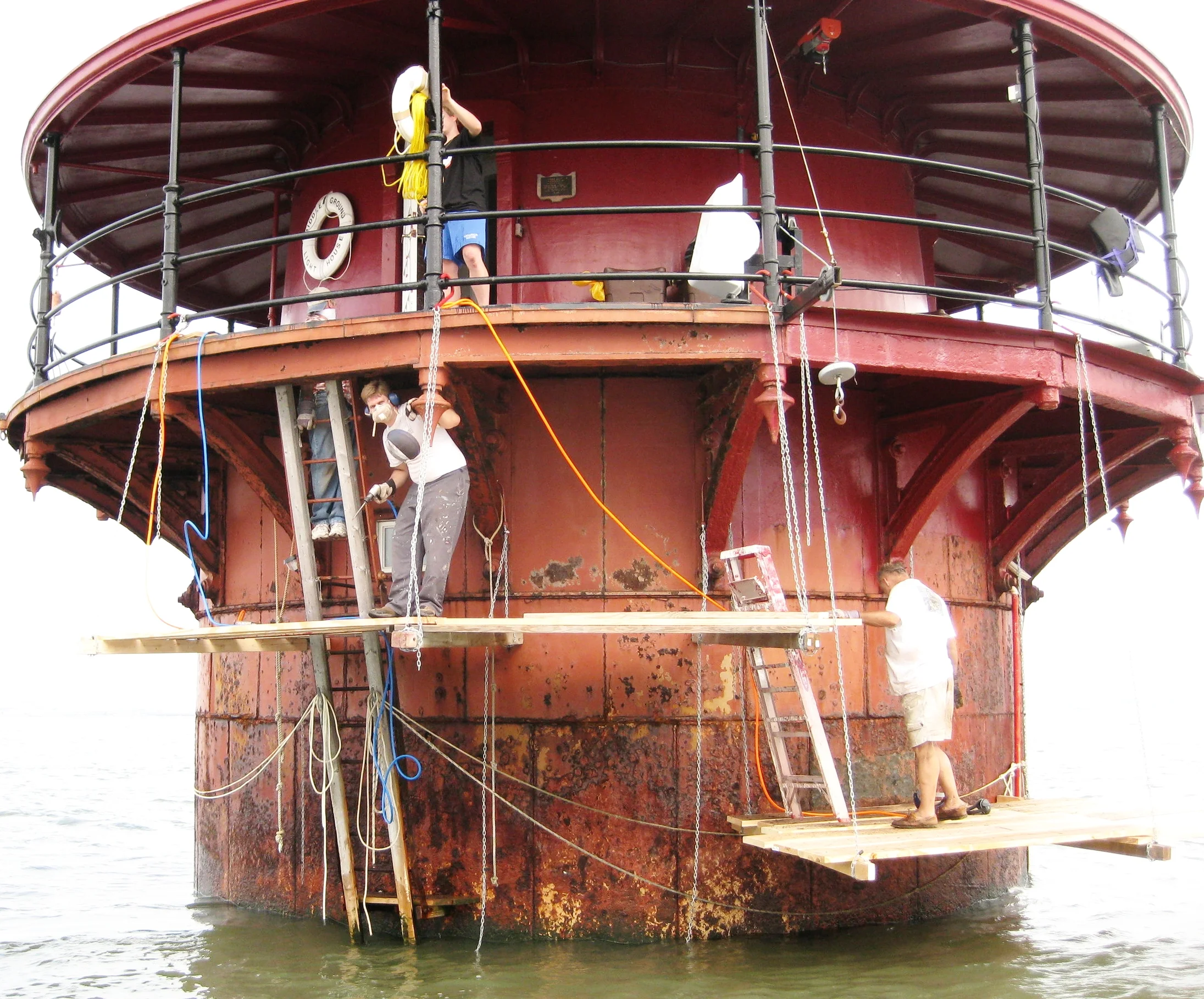 Workers repairing or maintaining the historic Middle Ground Lighthouse with ladders, ropes, and scaffolding around it.