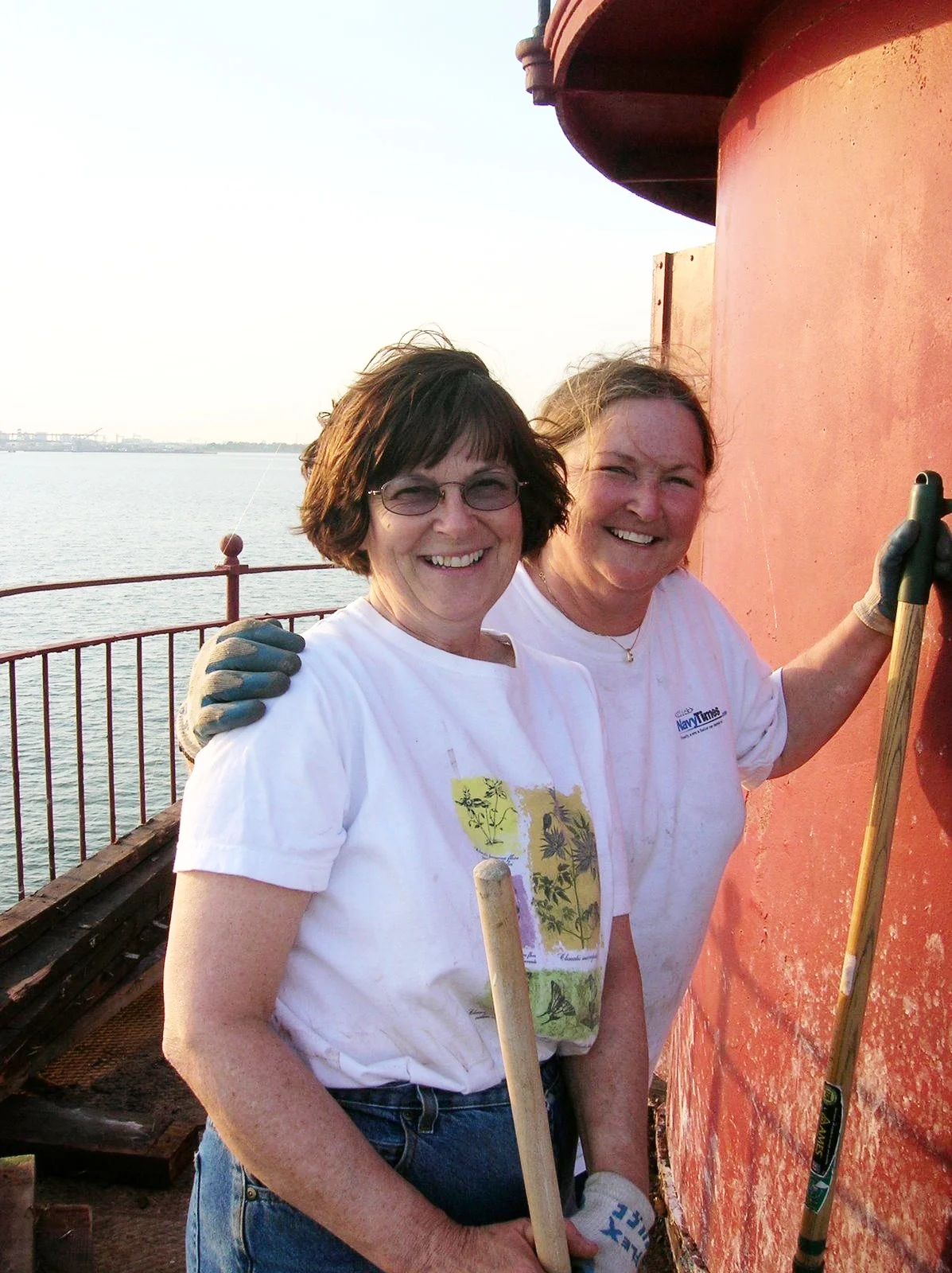 Workers on Middle Ground Lighthouse smiling at the camera, holding cleaning tools, with water and a distant shoreline in the background during sunset.