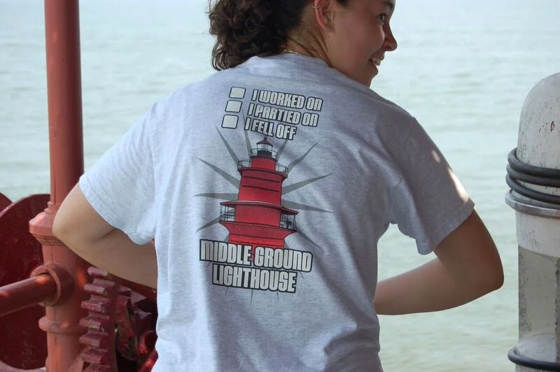 A woman wearing a gray T-shirt with a lighthouse graphic. The shirt has checkboxes with the options: 'Worked on,' 'Partied on,' and 'Fell off,' with 'Fell off' checked. The lighthouse is labeled 'Middle Ground Lighthouse.' The woman is on a boat or p