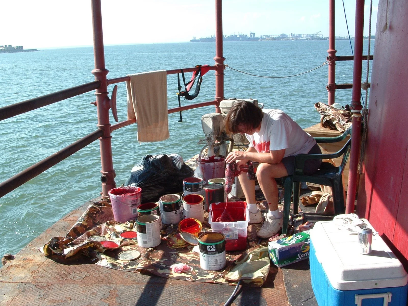 A person working on the main deck of Middle Ground Lighthouse with cans of red paint, paint supplies, and a cooler, while the ocean and distant ships of Hampton Roads are visible in the background. 