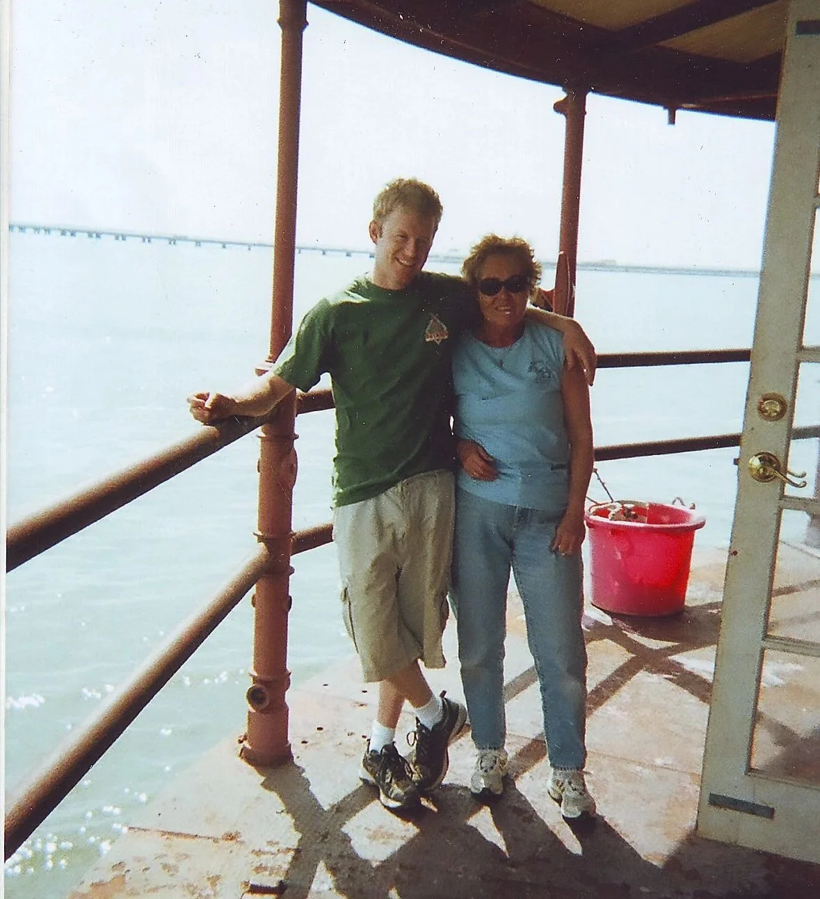 Two people, a young man and an older woman, are standing close together on Middle Ground Lighthouse, smiling, with the water and a bridge in the background. 