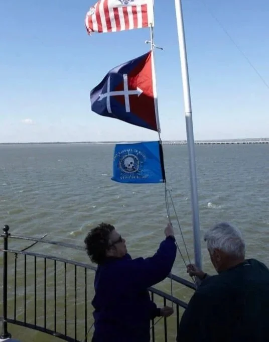 Two men raise a blue flag with a circular emblem at a waterfront, with an American flag and a red, white, and blue flag on a pole on Middle Ground Lighthouse. 