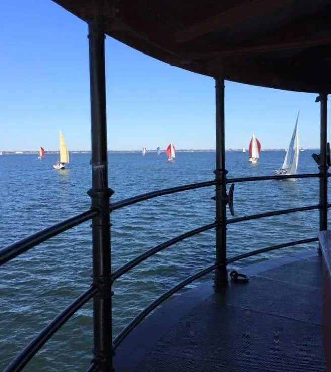 View of sailboats with colorful sails on a body of water, seen from Middle Ground Lighthouse with metal railing and canopy.