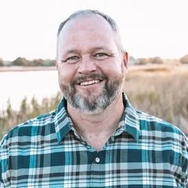 A middle-aged man with a beard and short hair smiling outdoors in front of a body of water and tall grass, wearing a plaid shirt.