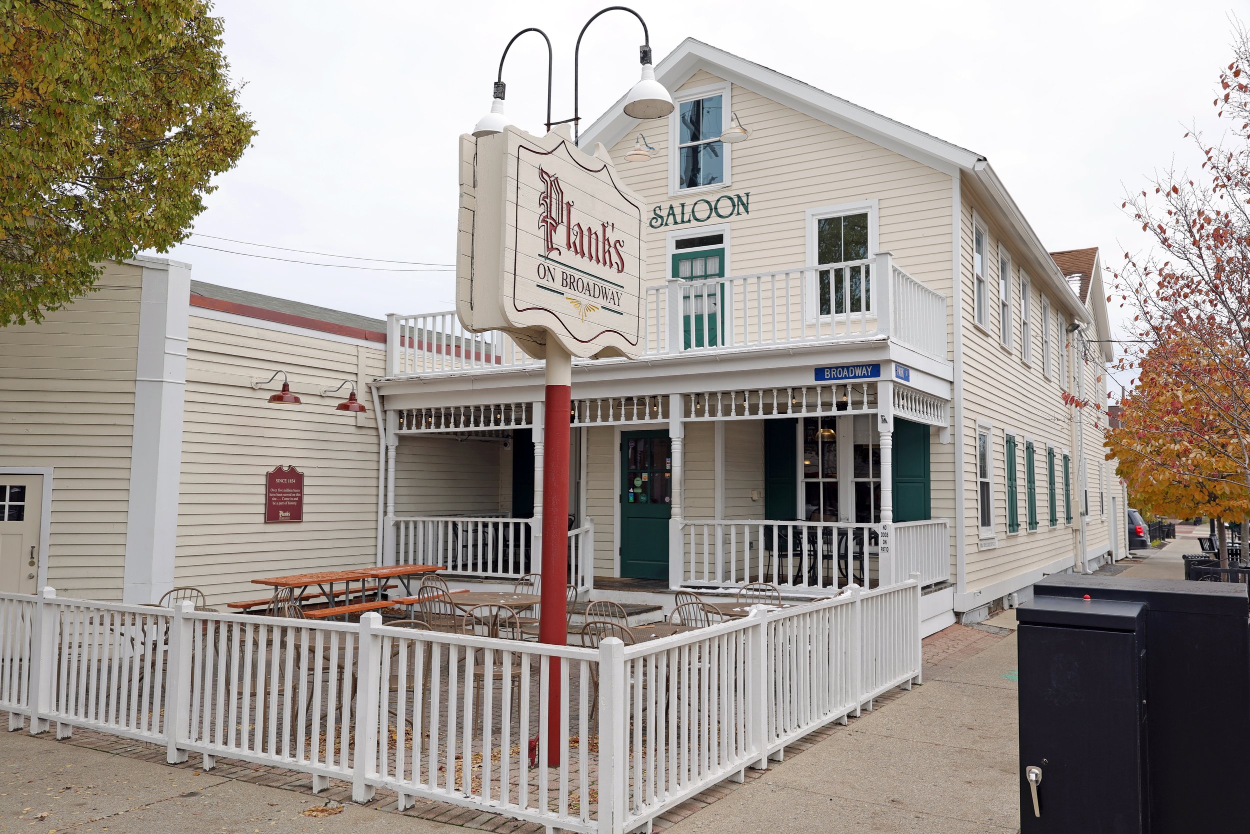 Exterior view of a two-story building with a sign that reads "Plank's on Broadway" The building has cream-colored siding, green shutters, and a porch with white railings. There is patio seating with tables and chairs in front.