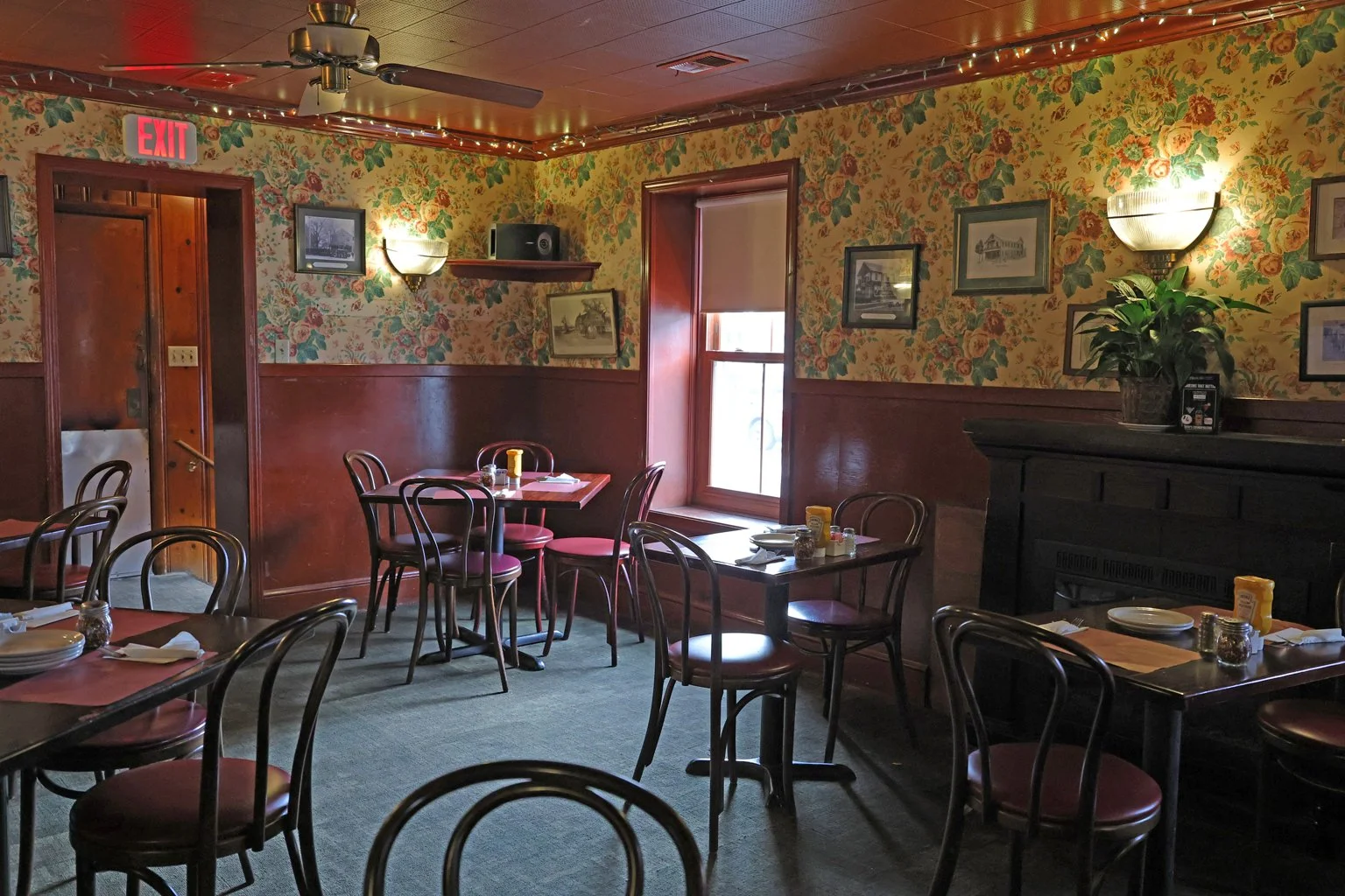 Interior of a cozy restaurant dining room with wooden tables and chairs, floral wallpaper, wall-mounted lights, and framed artwork.