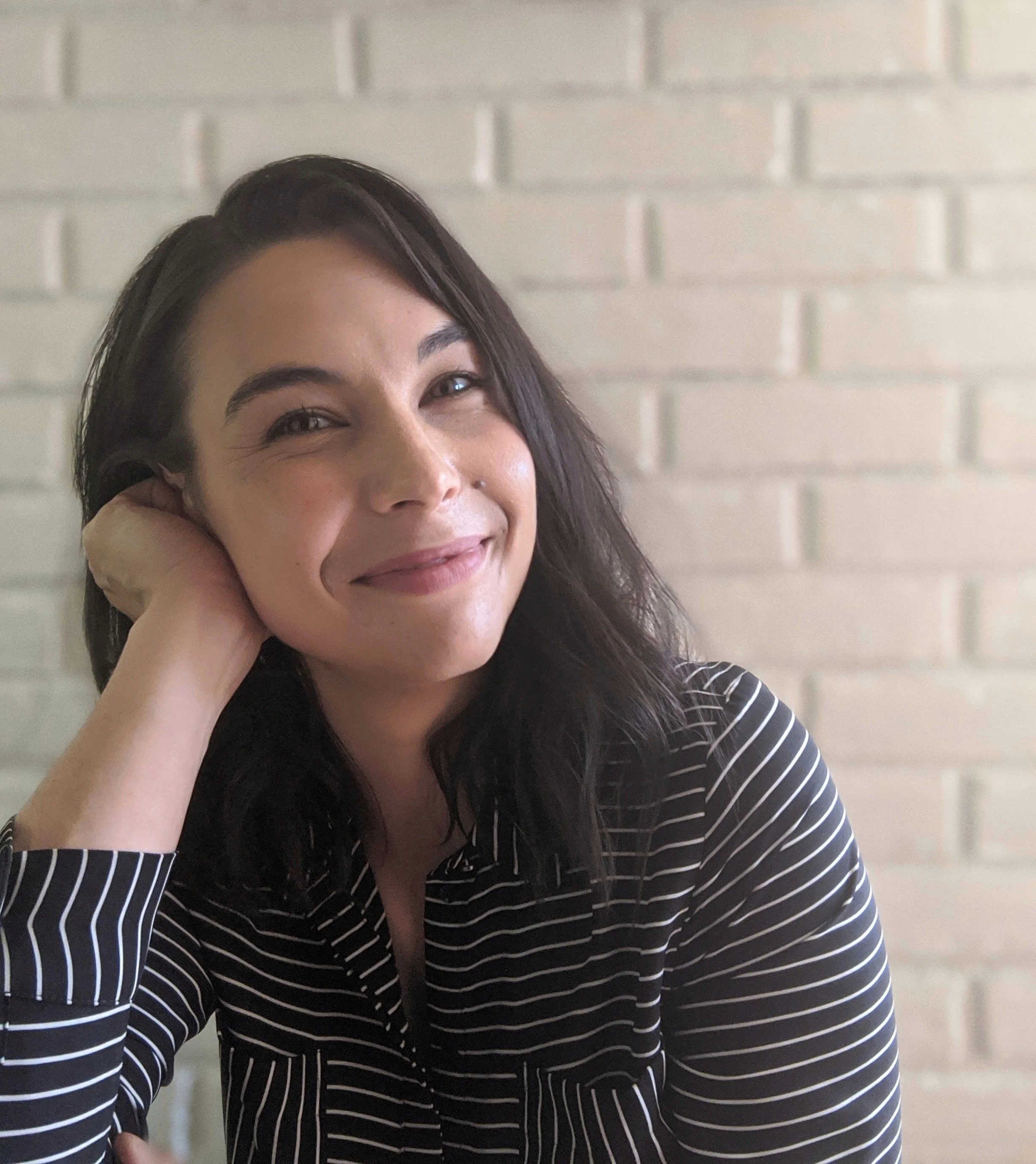 Rachel rests her head on her hand as she smiles. She has dark brown hair and wears a black and white striped shirt.