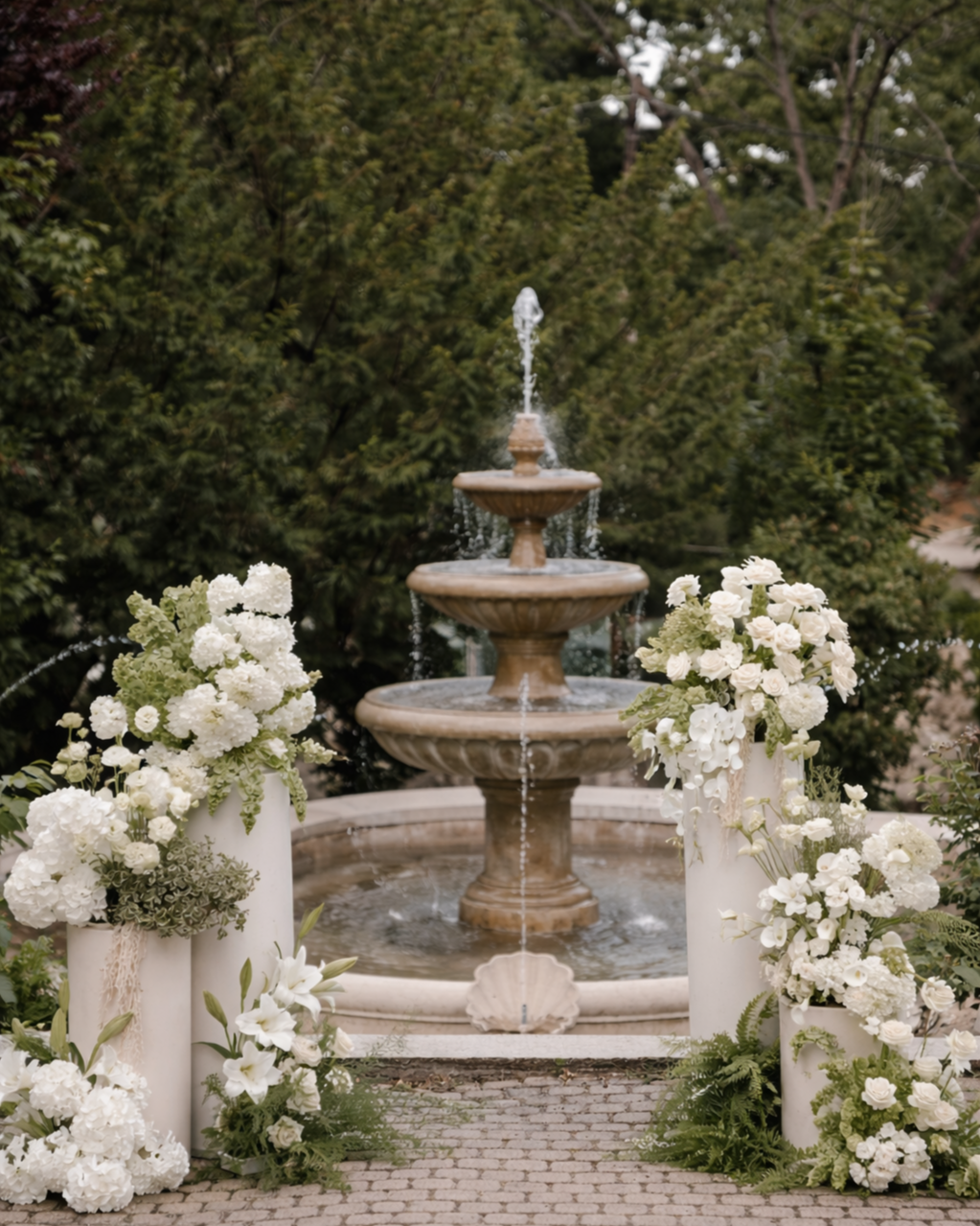 A decorative fountain with water cascading down three tiers, surrounded by white floral arrangements in large white vases, set on a cobblestone pathway with lush green trees in the background.