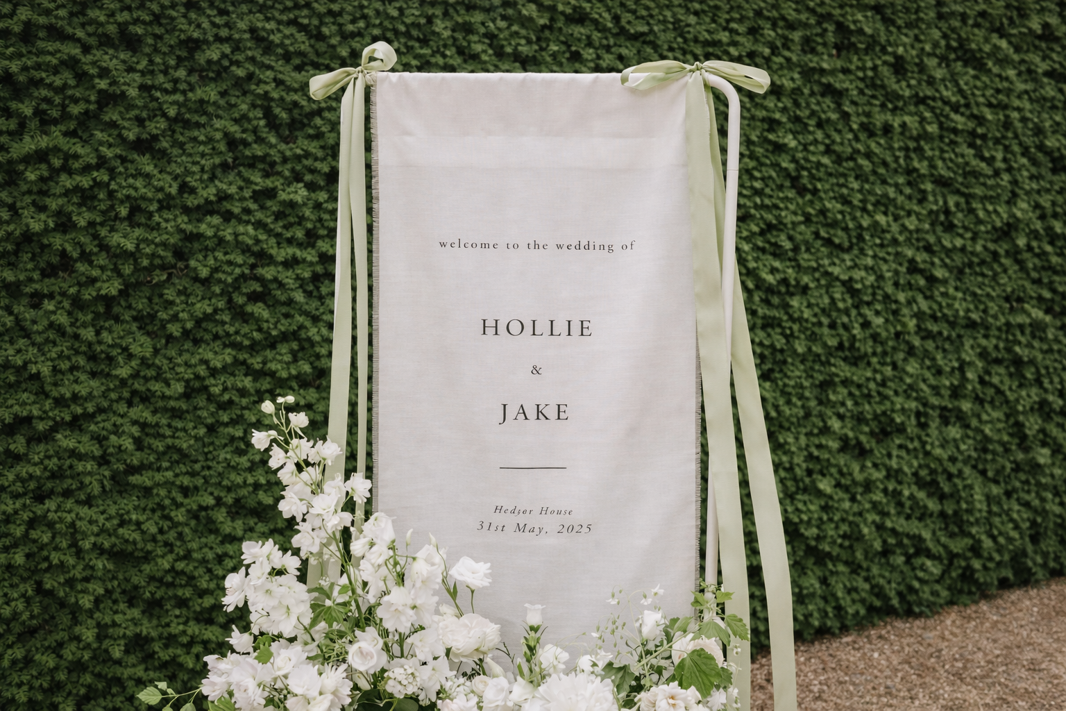 Wedding welcome sign with white flowers in front, green hedges in the background