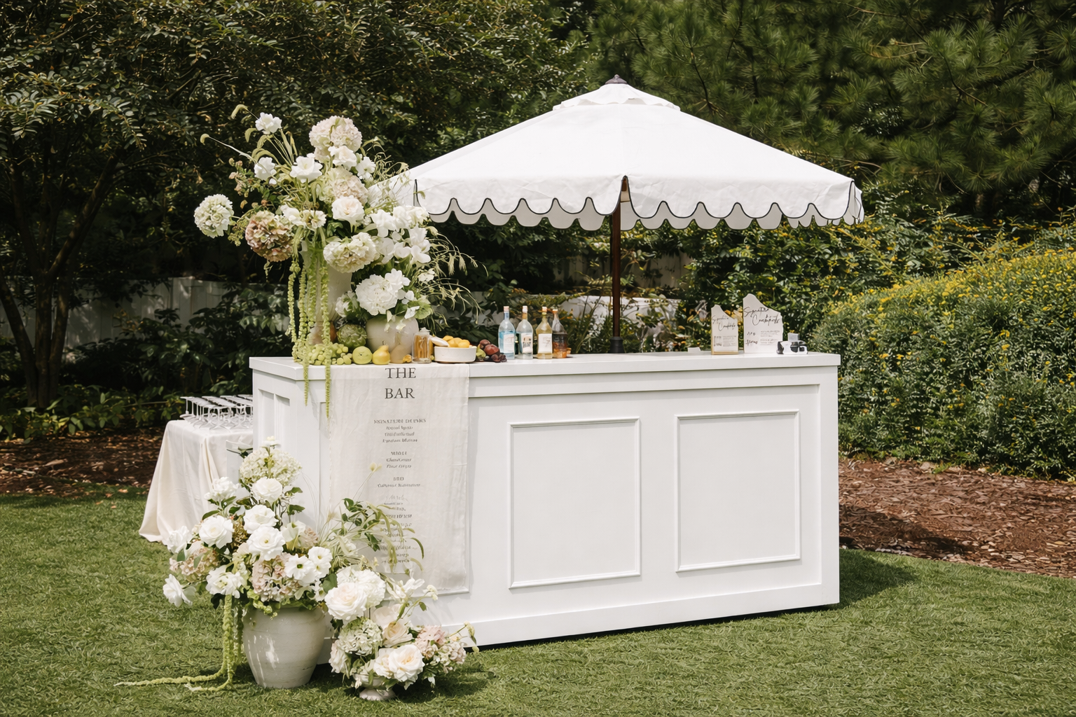 A white outdoor bar decorated with white and pastel flowers, set in a garden, with bottles of alcohol, glasses, and a menu, shaded by a white umbrella.