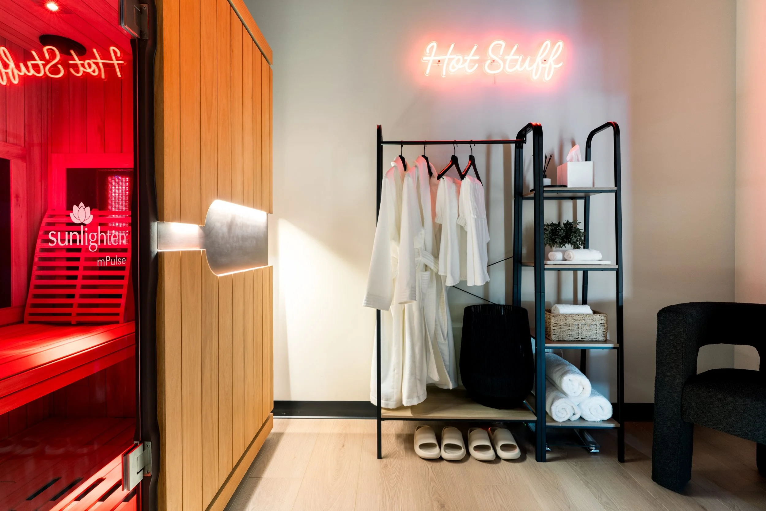 A spa or wellness room with a red hot stone sauna on the left, wooden walls, a clothing rack with white robes, a shelf with rolled towels, and a neon sign that says 'Hot Stuff' on the wall.