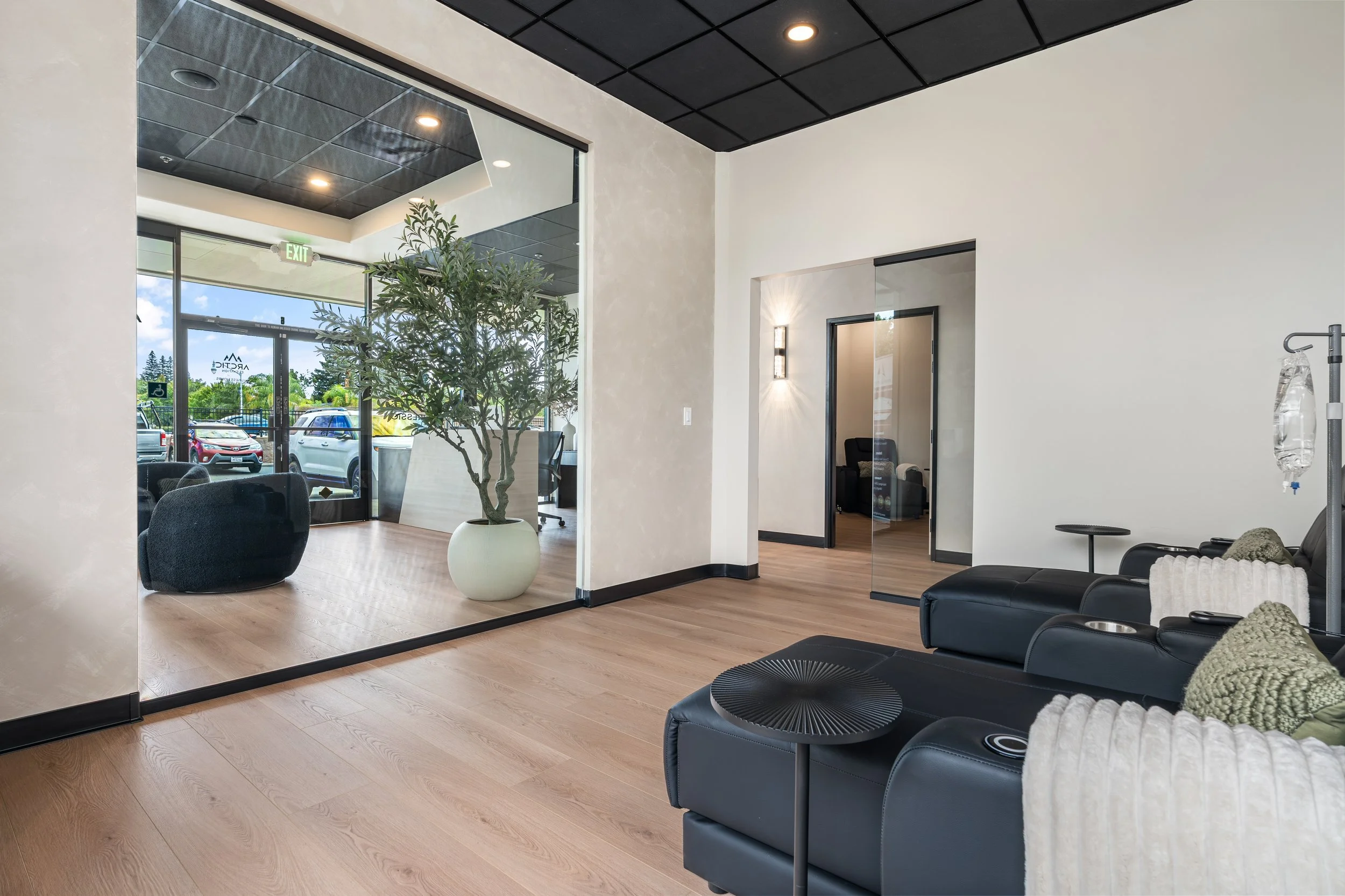 Interior of a modern waiting area with black leather seats, a small round black table, a potted plant, and large windows showing parked cars outside.