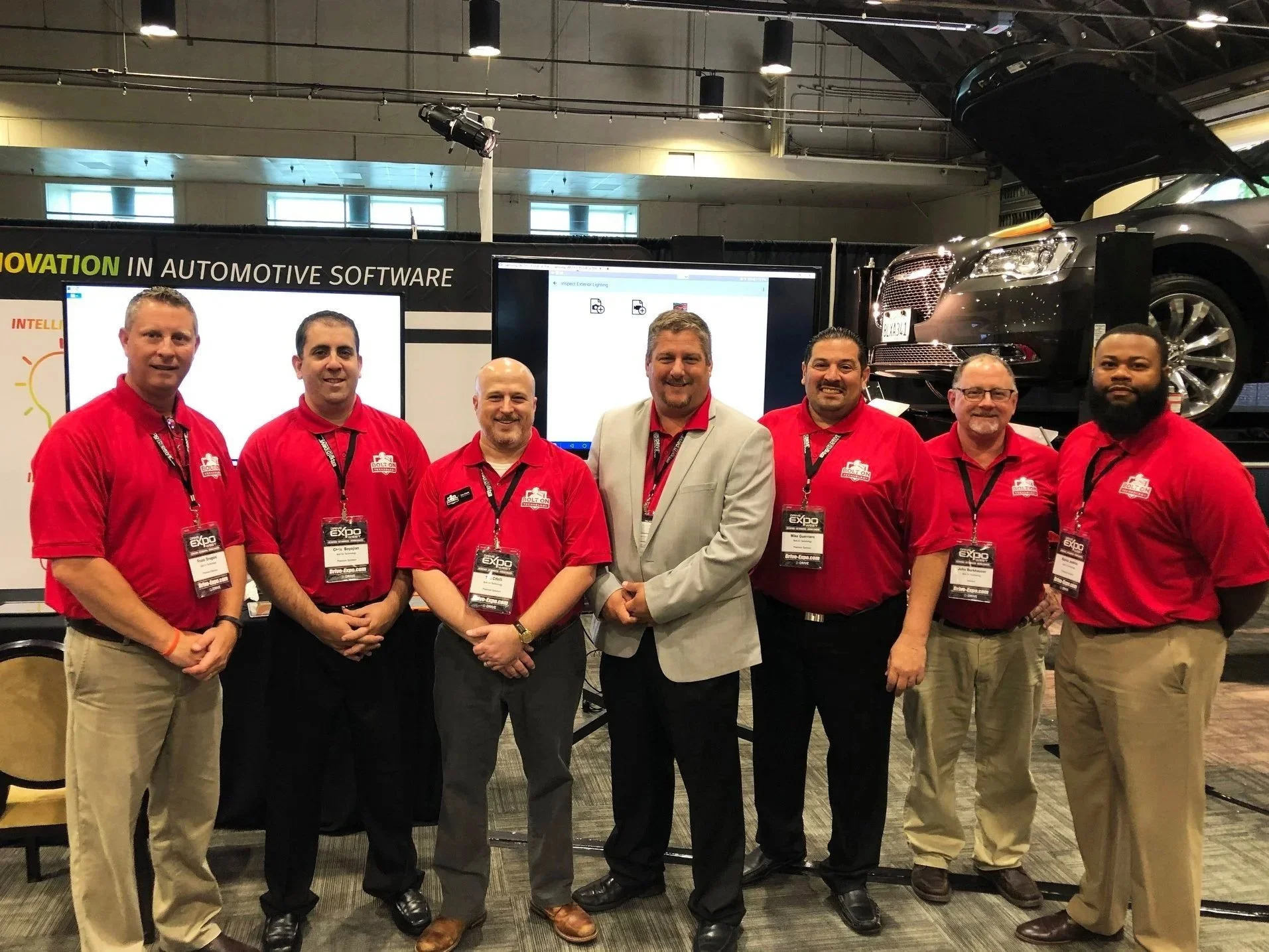 Group of seven men standing in front of an automotive technology display at a conference, with a car on a lift behind them. All are wearing conference badges, six in red shirts, one in a light gray blazer.