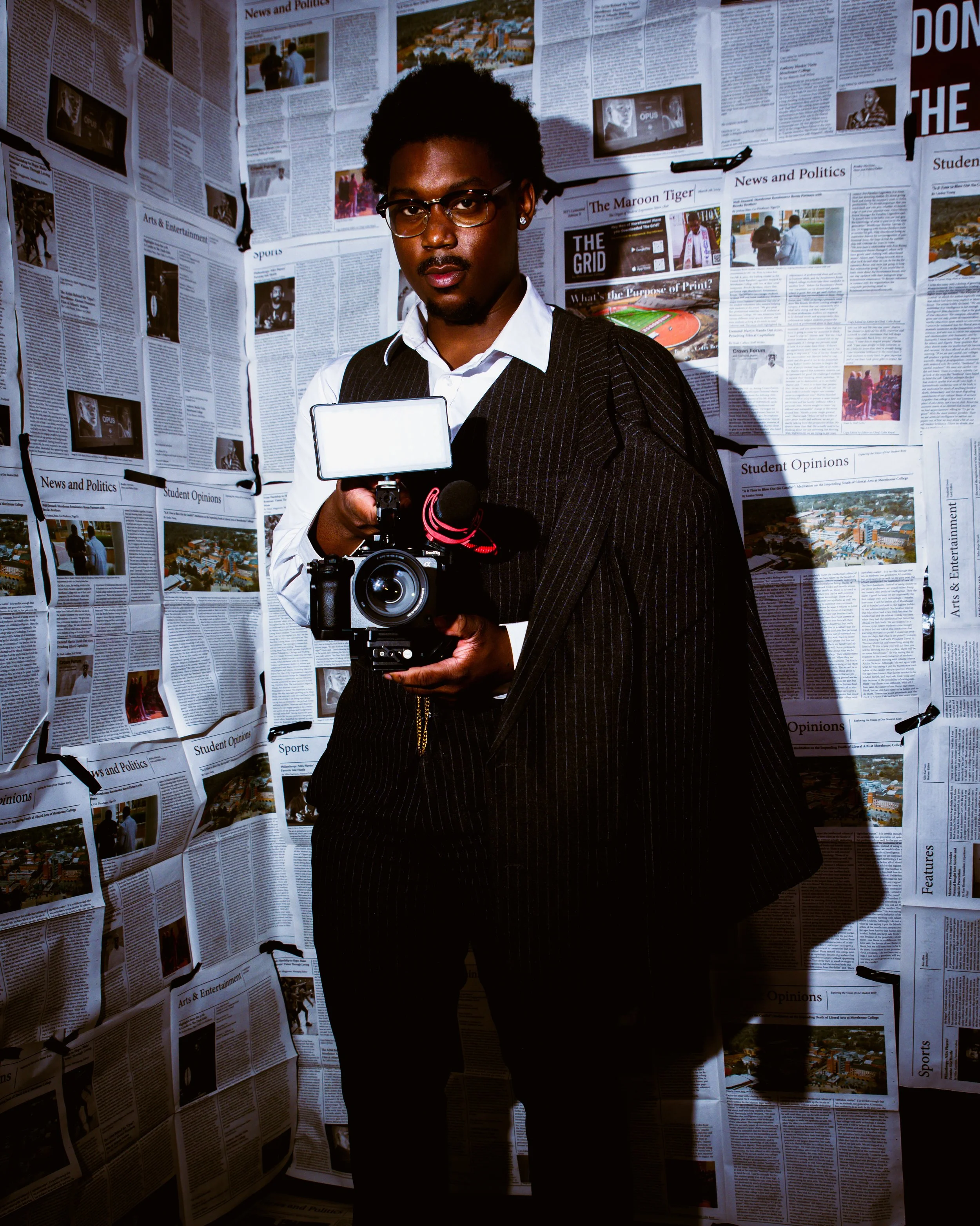 A man in a suit holding a professional camera with a light attached, standing in front of walls covered in newspaper pages.
