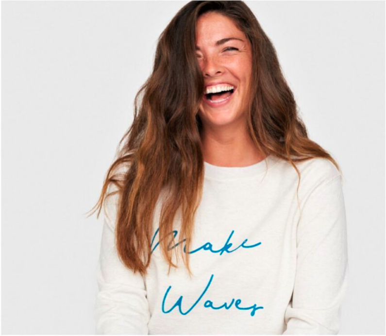 A woman laughing with long wavy brown hair, wearing a white sweatshirt with blue text that says "Make Waves", against a plain light background.