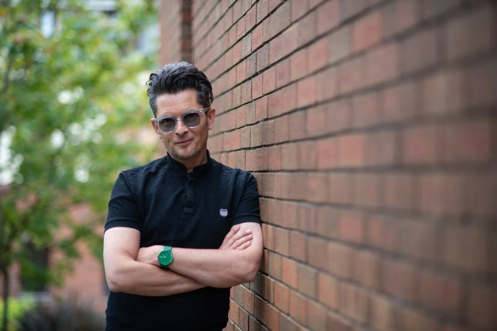 A man with dark hair wearing sunglasses and a black polo shirt, standing with arms crossed against a brick wall outdoors, with green trees in the background.
