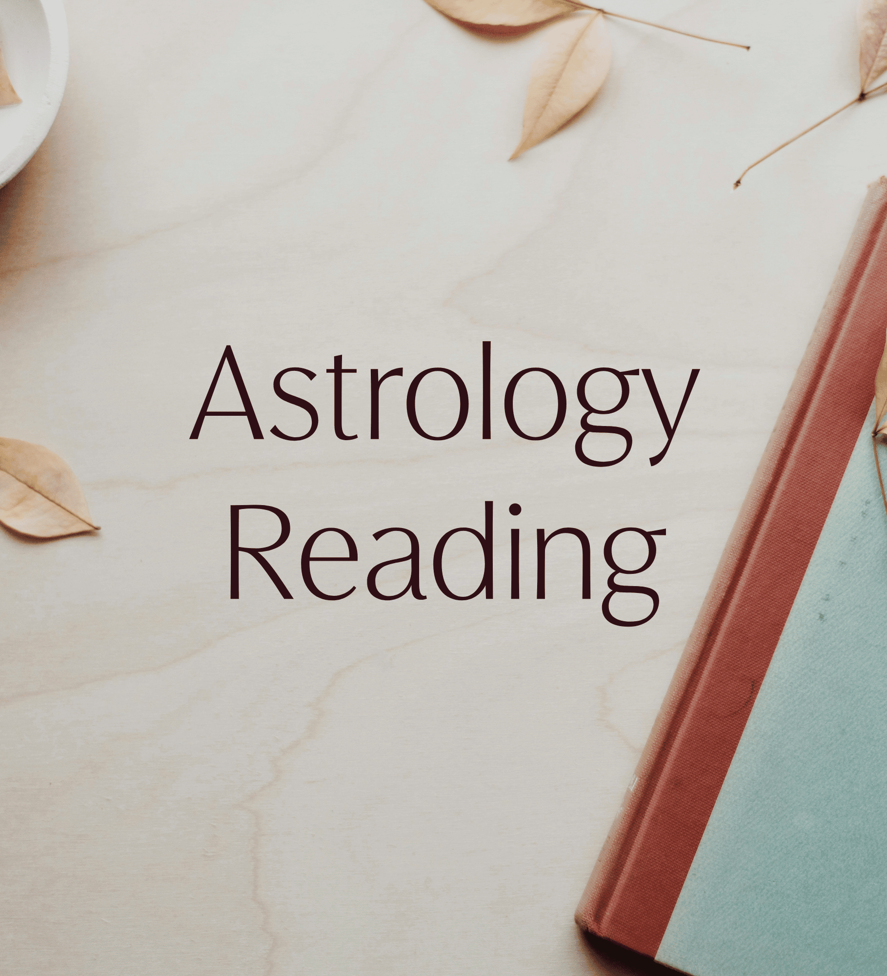 A close-up of a book titled 'Astrology Reading' resting on a light-colored wooden surface, with dried leaves scattered around and a book with a dusty pink cover partially visible on the right.