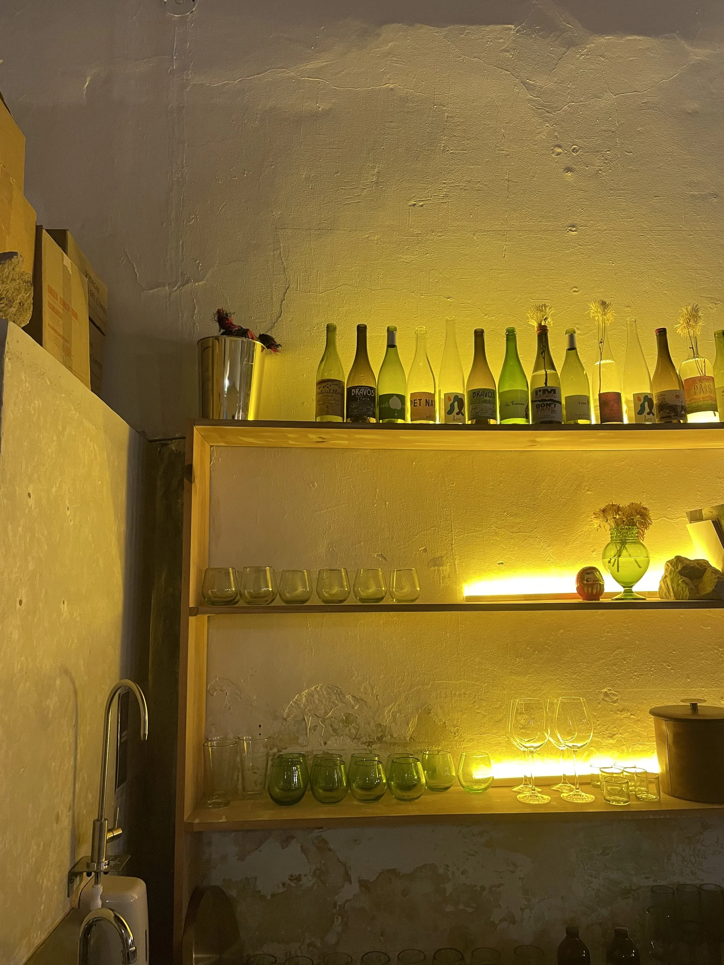 A wall-mounted wooden shelf with three levels holding glassware such as wine glasses, tumblers, and decorative bottles. The bottles are illuminated by warm yellow backlighting, creating a cozy ambiance. There is a small vase with flowers and some decorative rocks on the middle shelf. The wall behind is textured with peeling paint, and to the left, a stainless steel sink is partially visible.