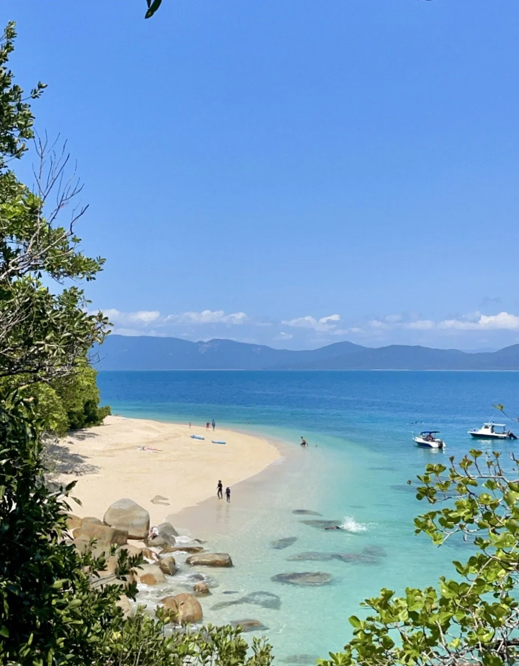 Beautiful beach with turquoise water, white sand, boats, and people, surrounded by greenery and distant mountains.